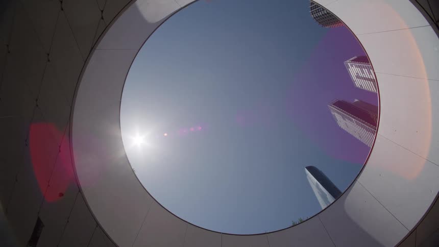 A slow pull back through a circular architectural opening reveals modern city skyscrapers against a sunny blue sky
