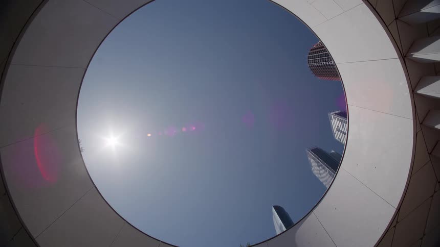 A slow zoom out from a low-angle shot reveals futuristic skyscrapers framed by a circular architectural opening against a bright, sunny sky