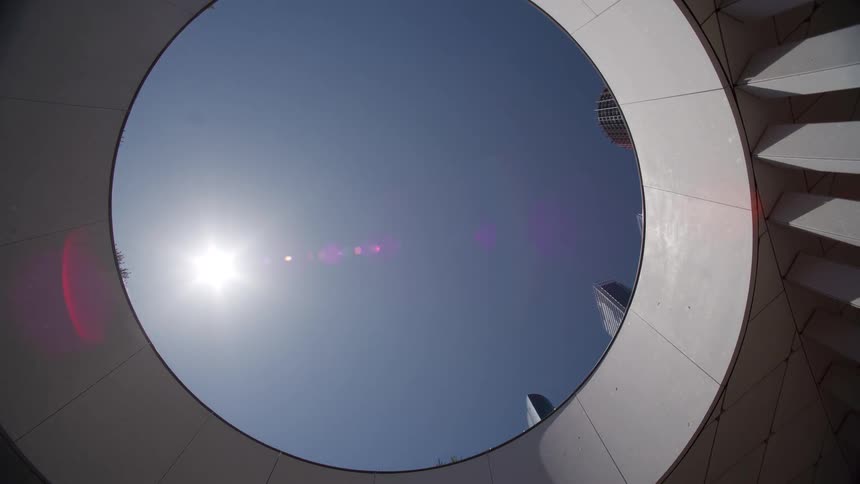 Looking up through a circular architectural feature, a slow pull-back reveals the bright sun in a clear blue sky, moving past modern skyscrapers