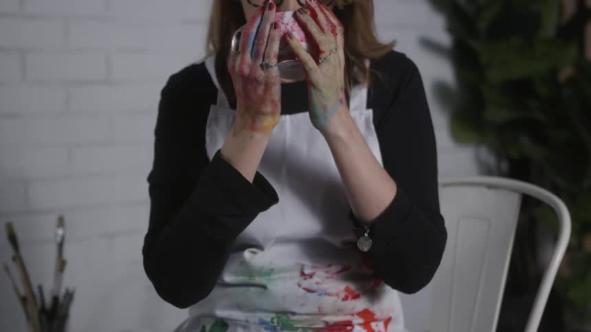 An artist is covered in paint and drinks from a mug