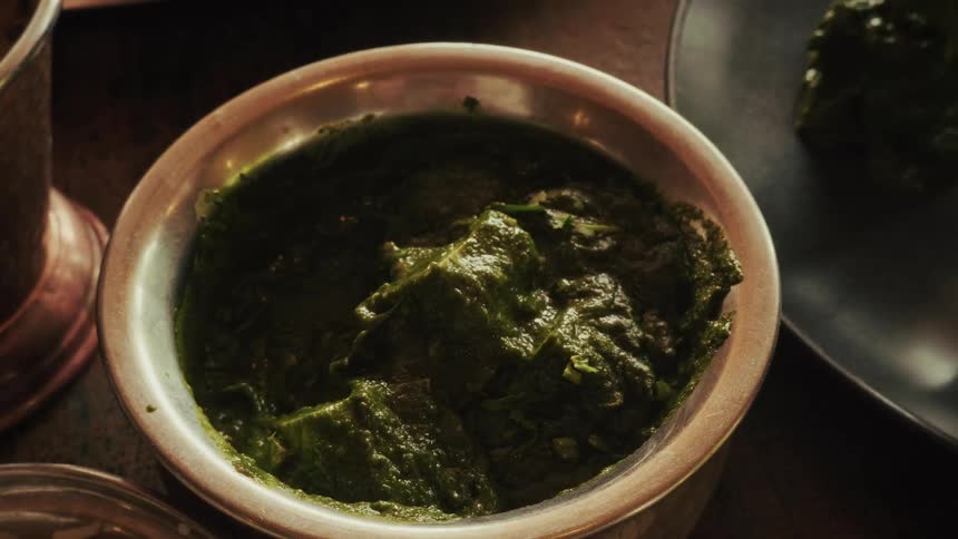 A close-up shot of a spoon scooping Palak Paneer from a traditional metal bowl at an Indian restaurant