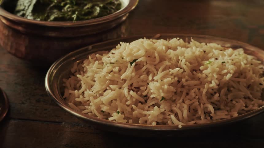 A slow zoom on a traditional metal bowl of delicious, steaming Indian basmati rice next to a serving of green curry