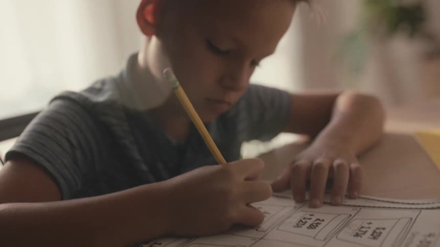 A focused young boy sits at a desk in a softly lit room, diligently completing his math homework with a pencil