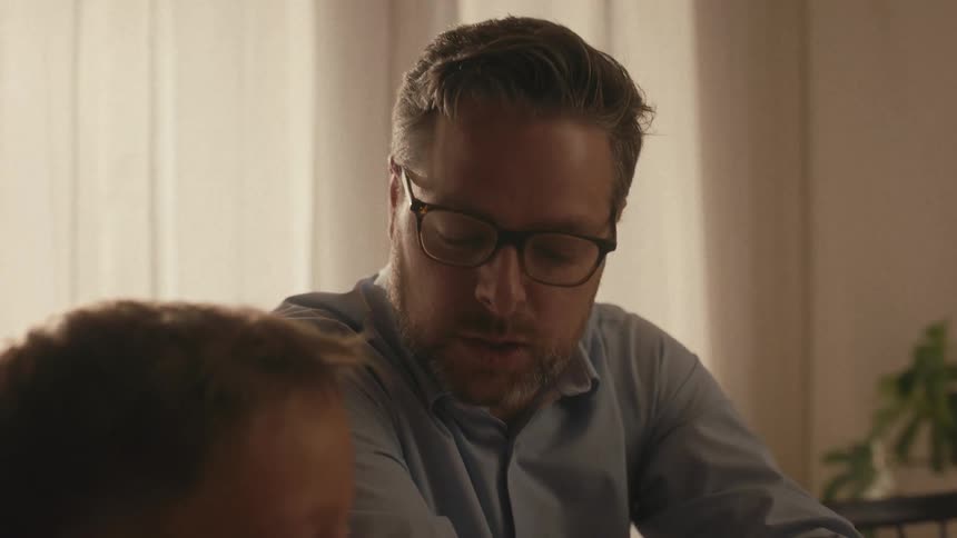 A caring father sits at the dining table helping his son and daughter with their homework in the evening