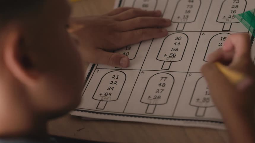 Close-up over the shoulder view of a young student diligently solving addition problems on a math worksheet with a pencil