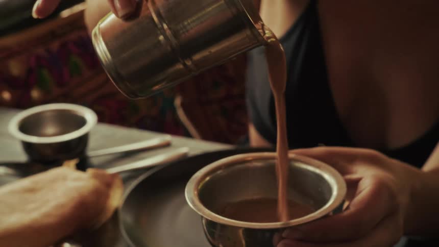 A person pours traditional Indian masala chai tea from one metal cup to another in slow motion at a restaurant table with authentic food