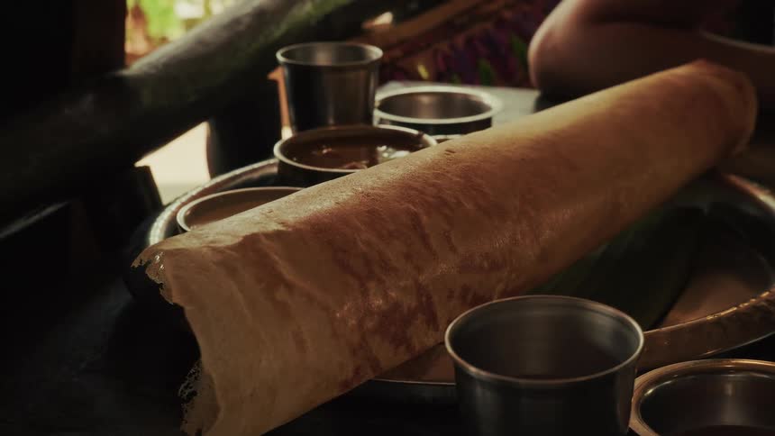 A delicious, crispy rolled dosa is served on a metal plate with various dipping sauces in a cozy restaurant