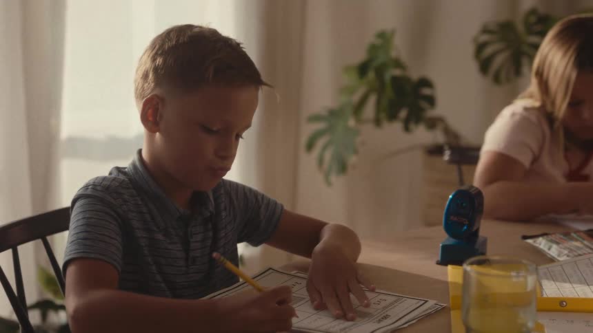 A brother and sister concentrate on their schoolwork at the dining table in their sunlit home