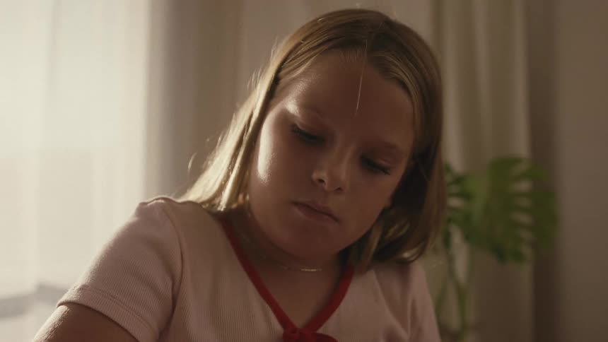 A young blonde girl concentrates as she uncaps a purple marker and begins to draw in a sunlit room