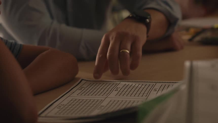 A patient father points to a math worksheet while helping his son with multiplication homework in slow motion