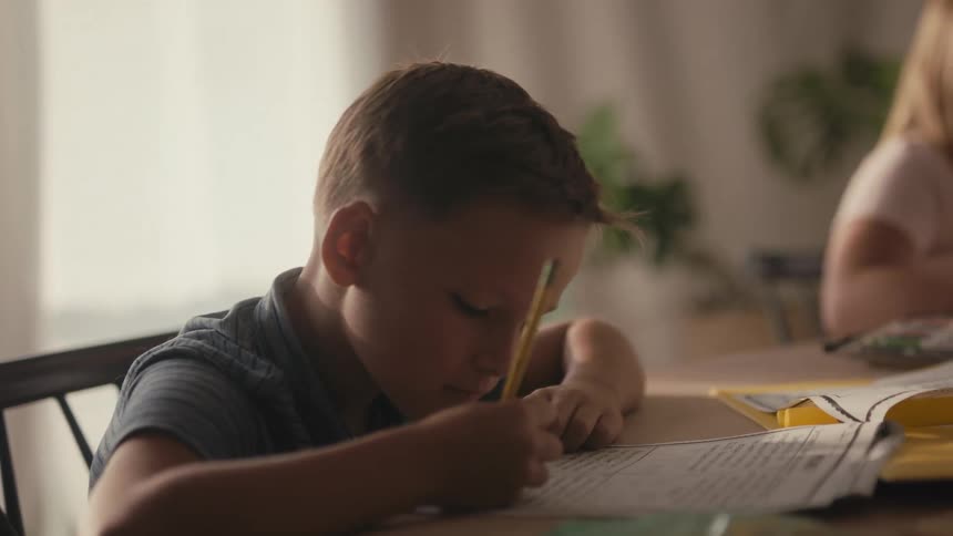 A young boy struggles to focus on his homework at the dining table, eventually resting his head in boredom as the shot reveals his sister also studying