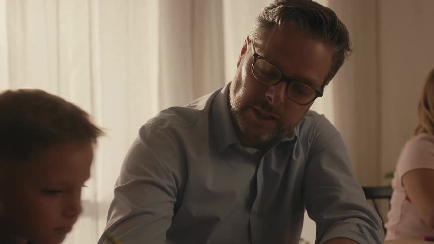 A caring father patiently helps his young son with homework at a table in a warmly lit room