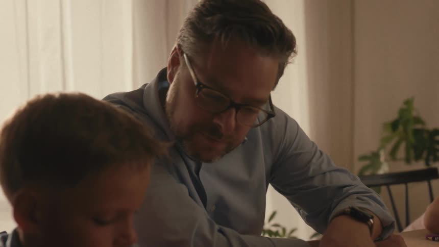 A caring father sits down to help his son who is concentrating on his math homework at the dining room table