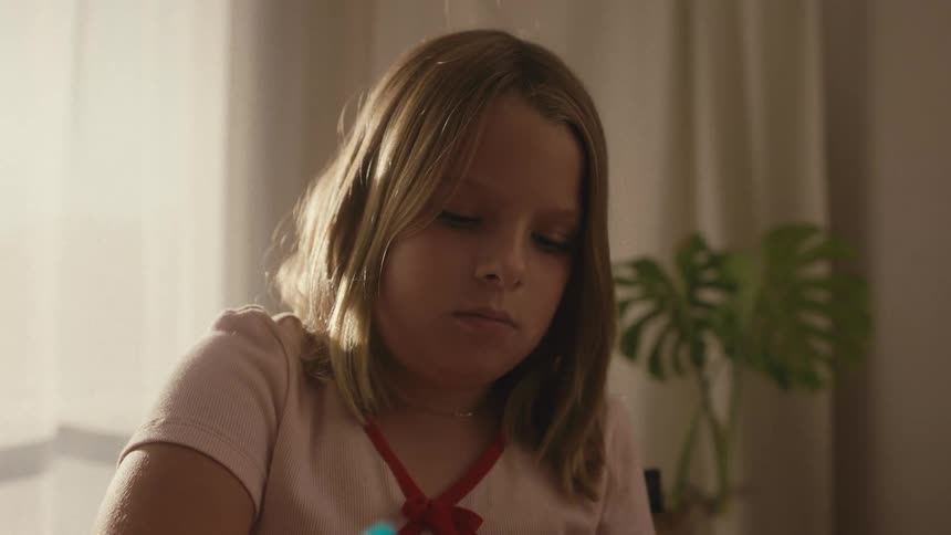 A focused young girl with blonde hair uncaps a blue marker to begin drawing in her room