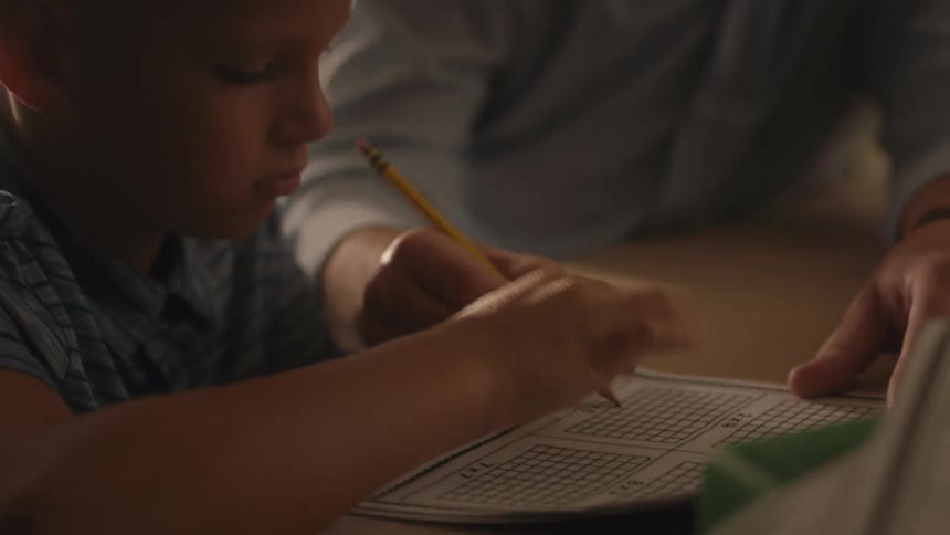 A close-up shot of a father patiently helping his young son with his homework at a table