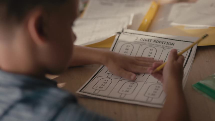 Over-the-shoulder shot of a young boy diligently working on his 2-digit addition math homework with a pencil
