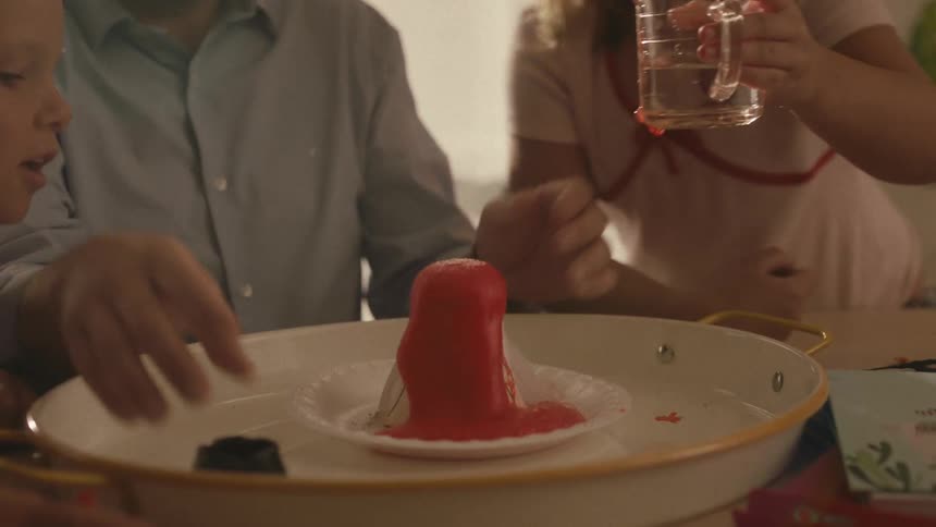 In slow motion, a father and his children watch in amazement as their homemade volcano science experiment erupts with red lava