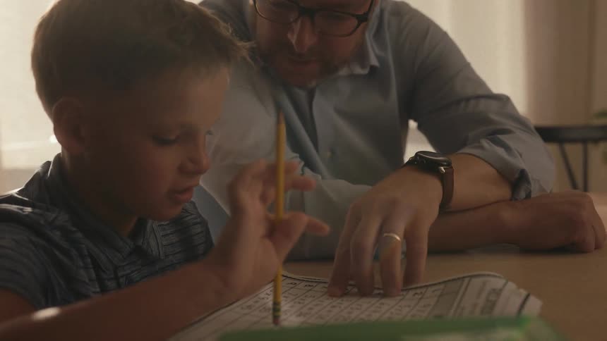 A patient father wearing glasses helps his young son with a math worksheet at a table in a warmly lit room