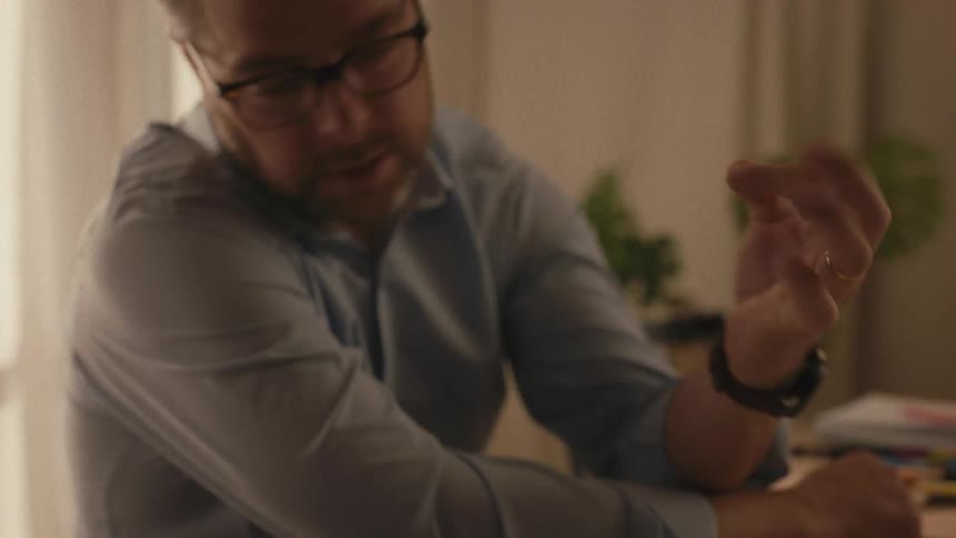A close-up shot reveals a father with glasses patiently explaining multiplication to his child using hand gestures over a worksheet