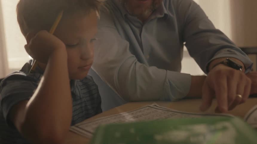 Patient father with glasses smiles as he helps his son with homework at a dimly lit table