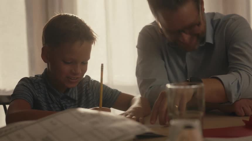 A caring father with glasses helps his young son with homework at a table in a warmly lit room