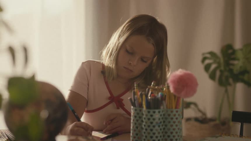 A slow rack focus from school supplies on a desk reveals a young girl diligently drawing in the afternoon light
