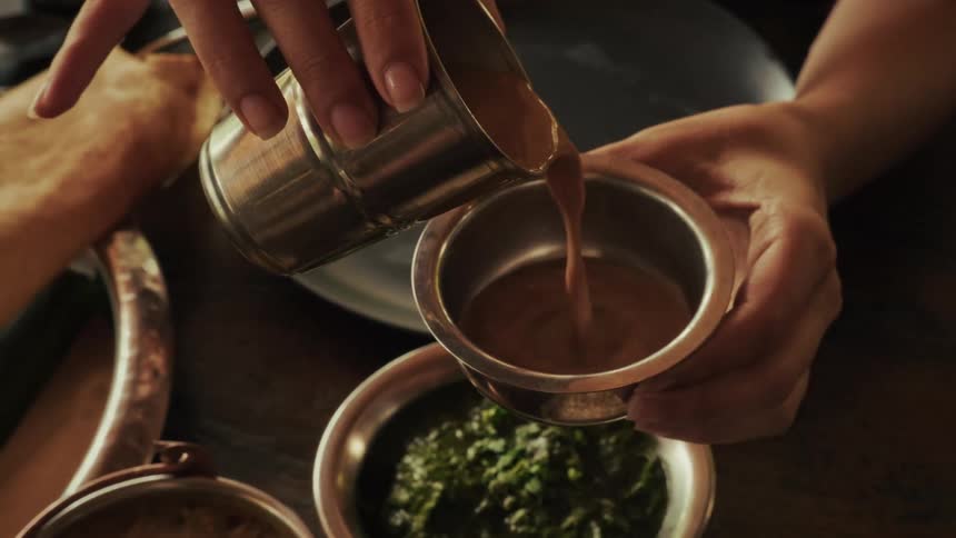 Pouring traditional Indian masala chai tea from a metal tumbler into a bowl in slow motion over a wooden table