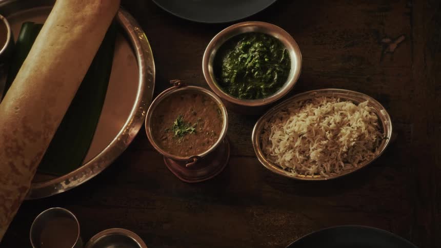 An overhead view shows a variety of traditional Indian dishes, including a dosa and dal, served on a dark wooden table