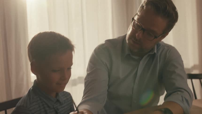 A caring father wearing glasses patiently helps his young son with homework at a sunlit table