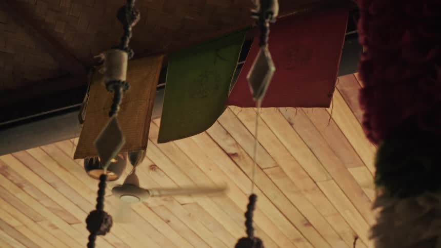 Decorative tassels and colorful prayer flags sway gently from a wooden ceiling while a fan spins