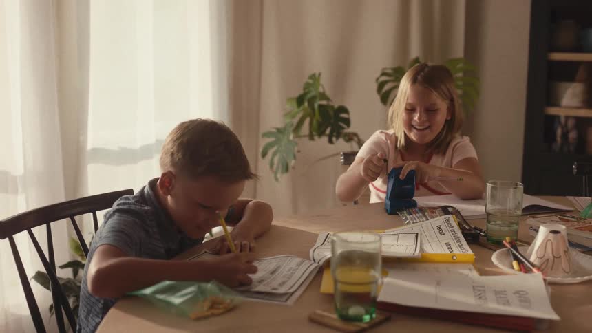 Happy brother and sister laugh while doing their homework together at the dining table in slow motion