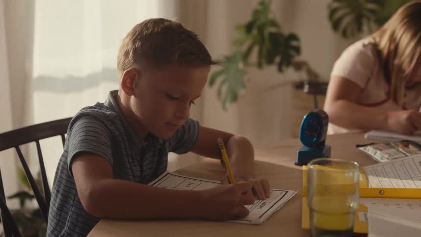 A young boy and his sister sit at the dining room table diligently working on their homework