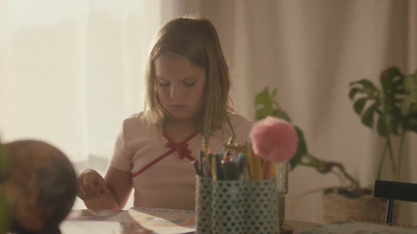 Rack focus from a potted plant to a young girl concentrating on her artwork at a sunlit desk