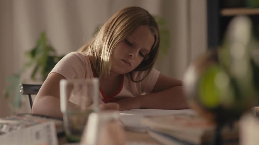 A young girl concentrates on her math homework at a cluttered desk in the warm light of her home