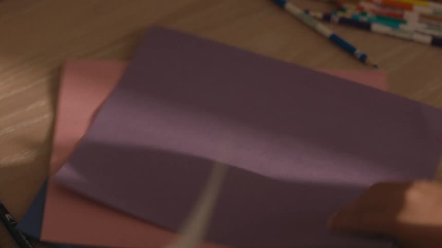 A child's hands place a blank sheet of white paper on a desk, ready to start drawing with colorful markers