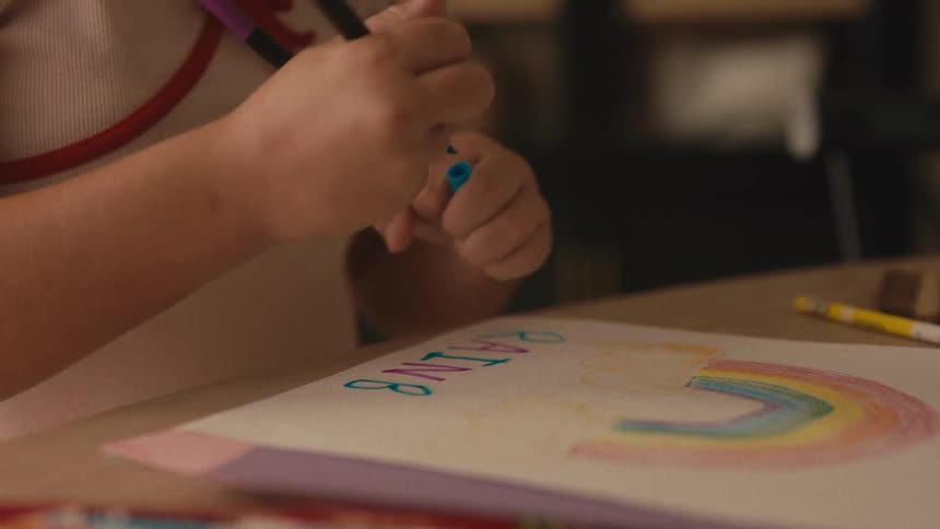 A close-up of a child's hands writing the word 'rainbow' in colorful letters next to a drawing of a rainbow