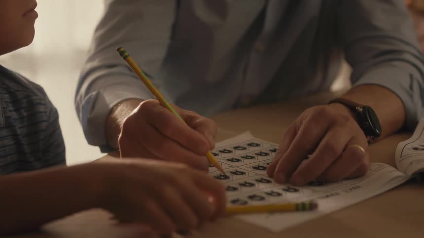 A close-up shot tilts down from a young boy's focused face to his father's hands guiding him through a homework worksheet with a pencil