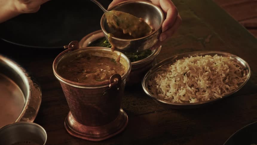 A woman slowly scoops rich and creamy Indian lentil dal into a small silver bowl at a rustic dinner table