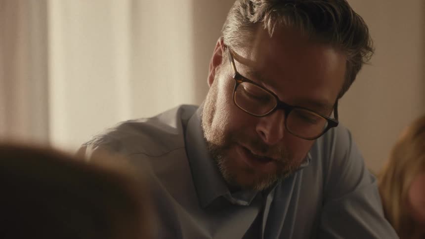 A father wearing glasses patiently guides his young son's hand as they work on a math worksheet together at home