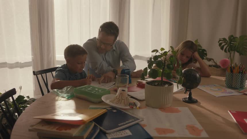 A supportive father helps his son with homework at the dining table while his sister looks on