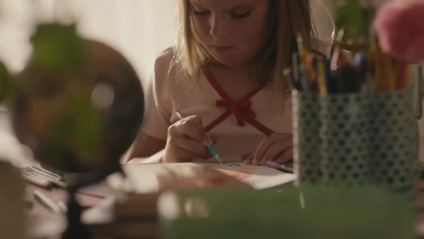 A young girl thoughtfully concentrates on drawing and coloring a rainbow at her desk in a warmly lit room