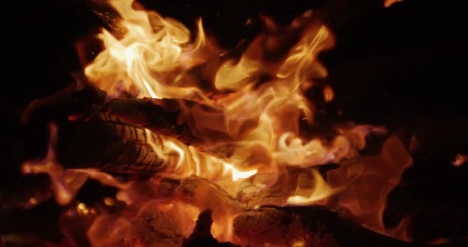 A close-up shot captures the mesmerizing dance of flames in slow motion as a campfire crackles and glows against a dark night