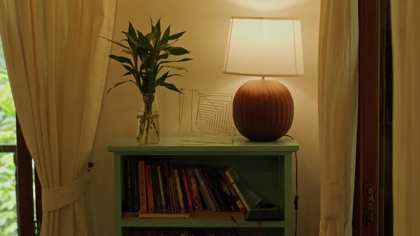 A cozy and warmly lit reading nook with a bookshelf, table lamp, and bamboo plant