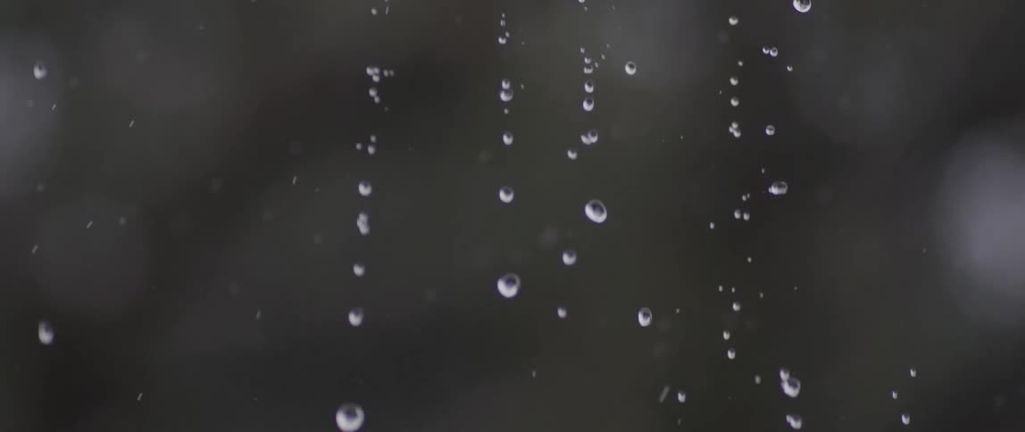 Raindrops fall in extreme slow motion from an overhang against a dark, blurry background