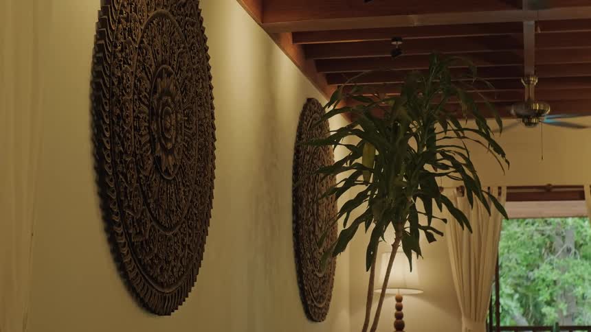 A serene room with Thai-style carved wooden art on the wall, a houseplant, and a ceiling fan