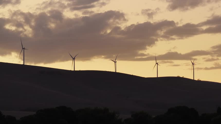 Silhouetted wind turbines on a rolling hillside generate power against a warm, cloudy sunset sky