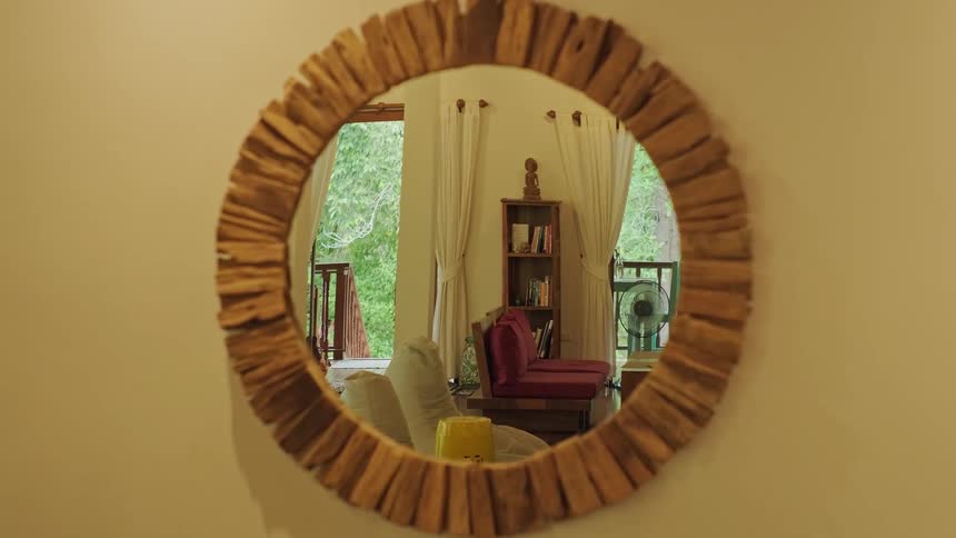 A rustic wooden mirror reflects a calm and cozy living room with a view of lush greenery outside