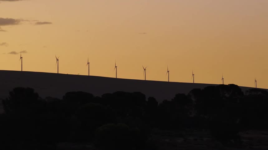 Silhouetted wind turbines spin slowly on a rolling hill against a golden sunset sky as a car drives in the foreground