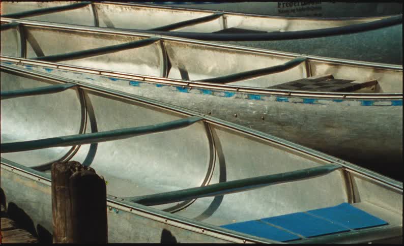 A close-up static shot shows sunlight reflecting off several empty aluminum canoes docked together