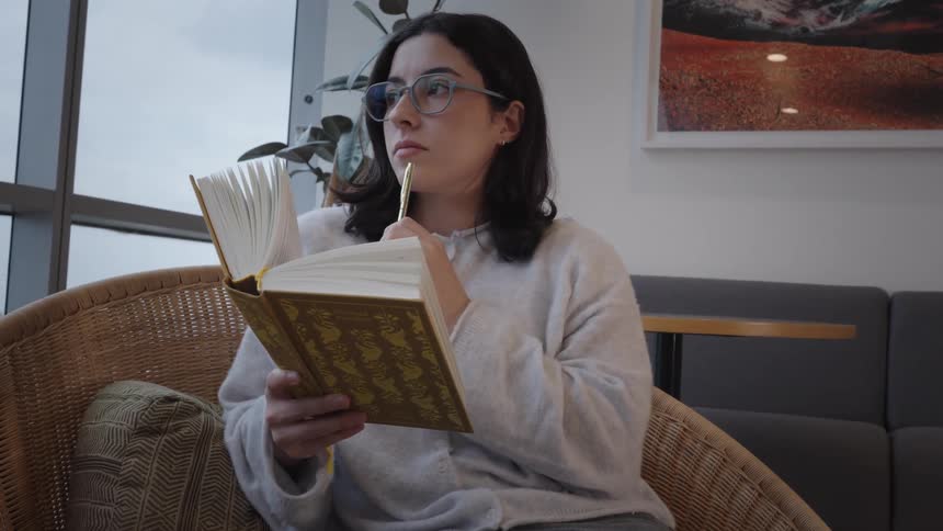 A thoughtful young woman wearing glasses reads a book and pauses to reflect while sitting in a comfortable chair by a window